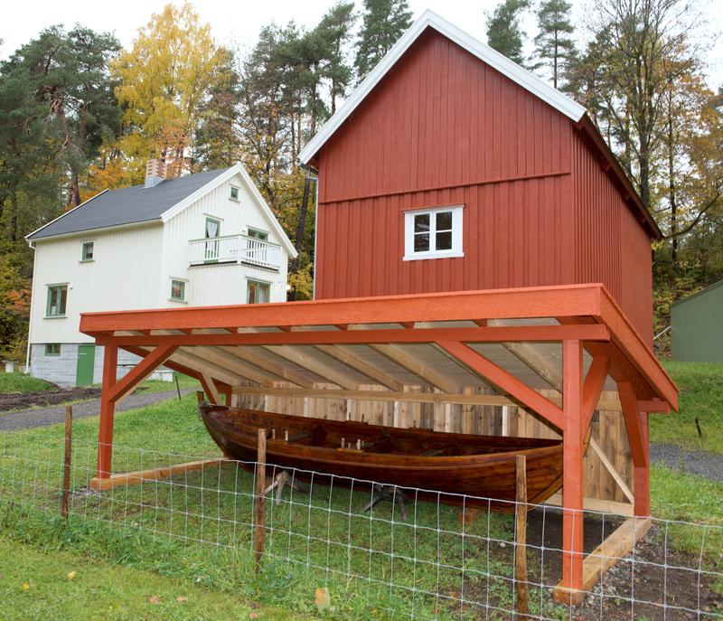 En båt ligger under et overbygg. Et bolighus og et fjøs i bakgrunnen.