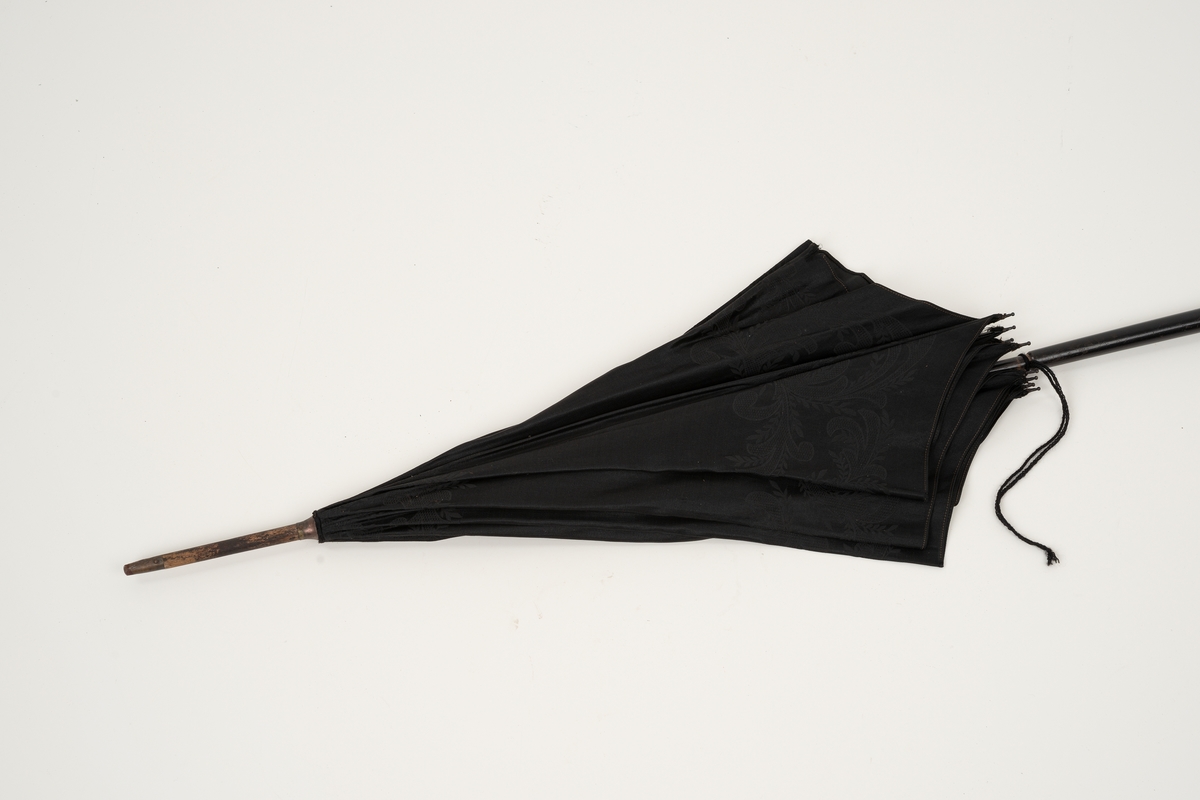 Svart parasoll med treskaft. Utskjæringer nederst i skaftet. Åtte metallspiler, trukket i svart silkedamask med organisk mønster.