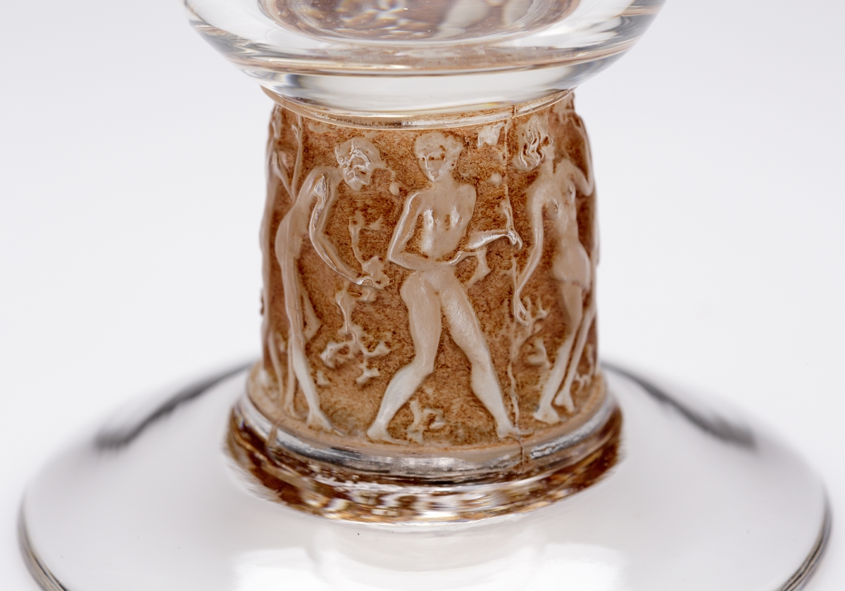 Vinglas med kupa i klarglas och ben dekorerat med kvinnofigurer. Runt reliefmönstret är glaset bruntonat.