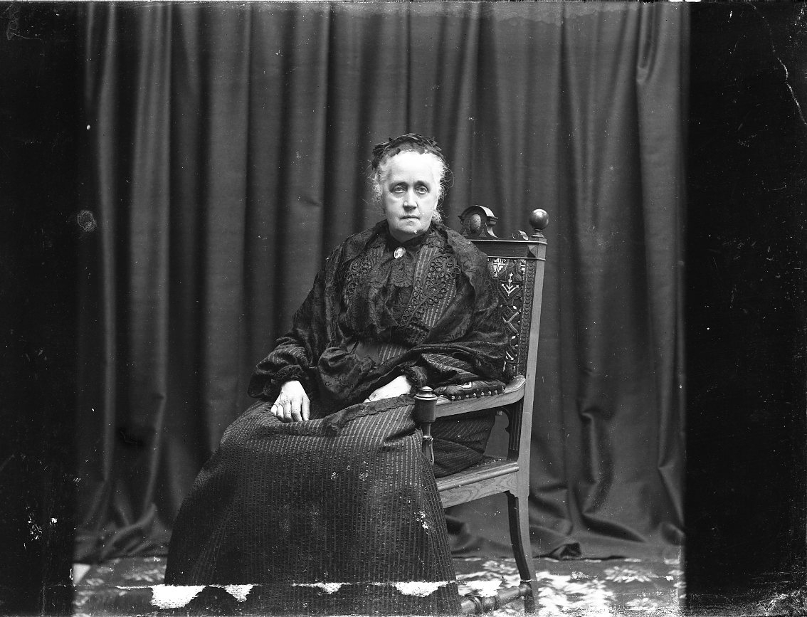 Porträtt av friherrinnan von Düben. Hon sitter på en stol i mörk dräkt framför en mörk drapering.