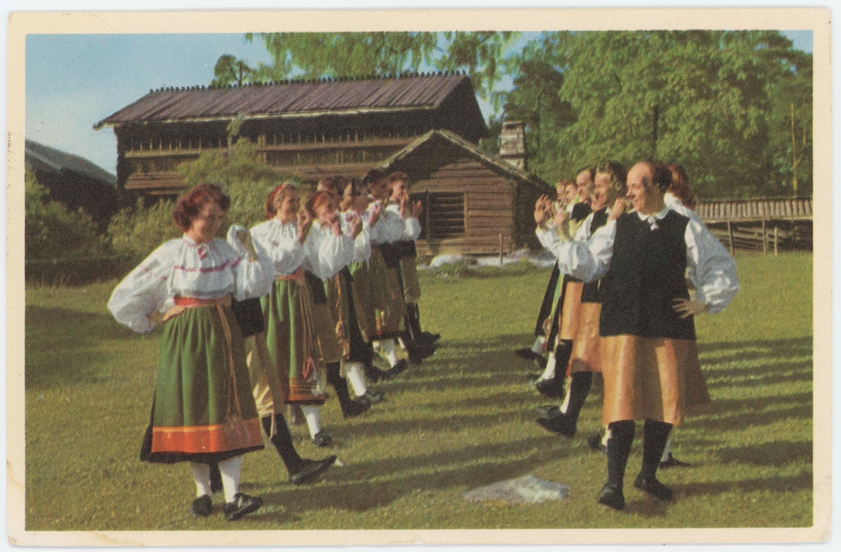 Vykort med motiv från Skansen. "Folkdanslag från Orsa, Dalarna". I bakgrunden "Orsastugan".