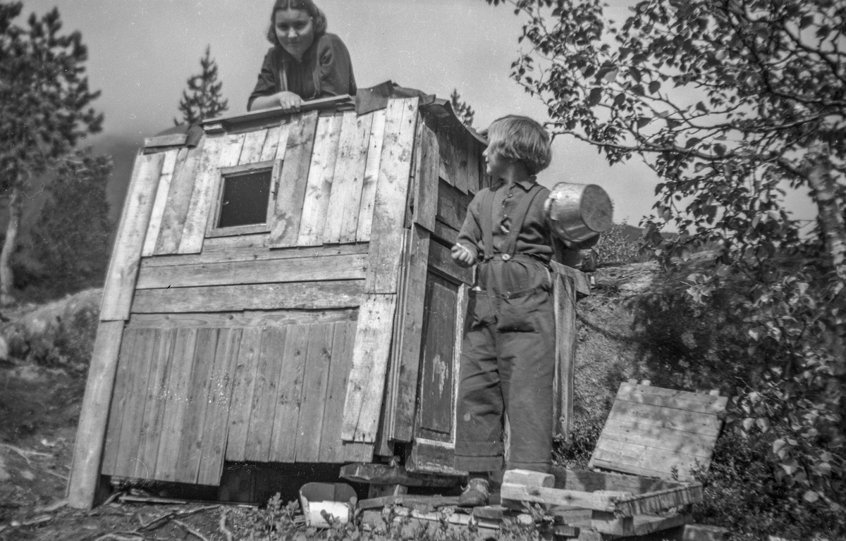To jenter ved leikehytte bygd av kassefjøler. 