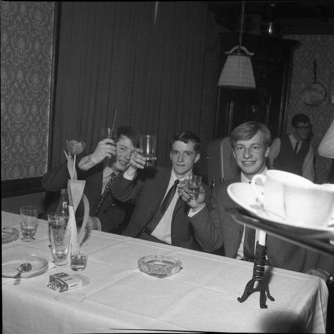 Tre unga och glada män i kostym och slips sitter vid ett bord och lyfter sina (grogglas) mot fotografen. På bordet står det flera tomma glas, askkopp, cigaretter och några använda assietter. En bricka med kaffekoppar kommer in från höger. Fotografi i samband med årsmöte för Gränna AIS.