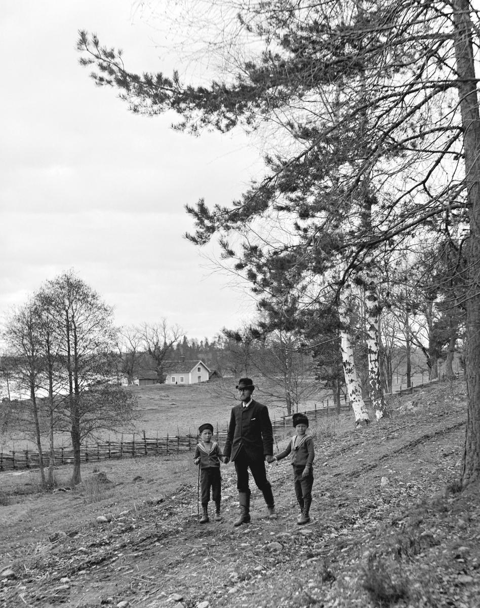 Karl Johan Hallin på promenad med sina söner Uno i vänstra hand och Sigurd i hans högra. I bakgrunden syns torpet Middagsfisket under Ramsdal.