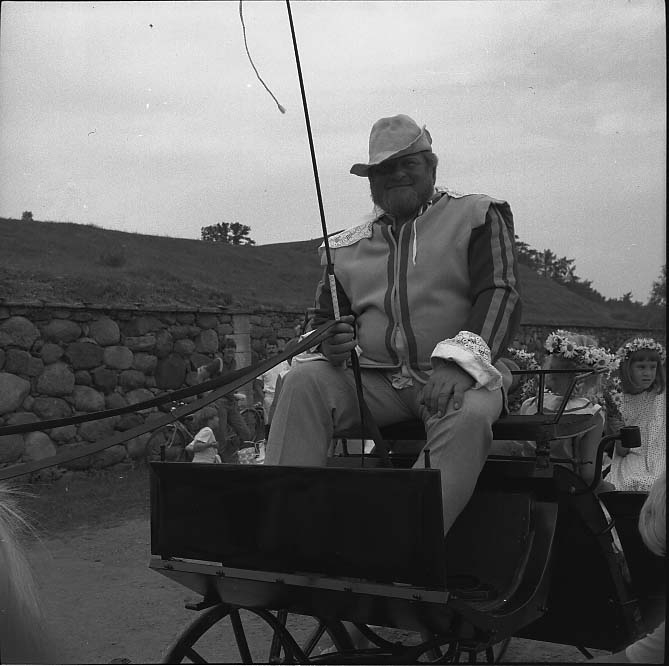 Lars Roempke sitter i historisk dräkt på kuskbocken till en vagn nedanför Visingsborgs slottsruin på Visingsö. Flickor i ljusa klänningar och blomsterkransar på huvudet sitter i vagnen.
