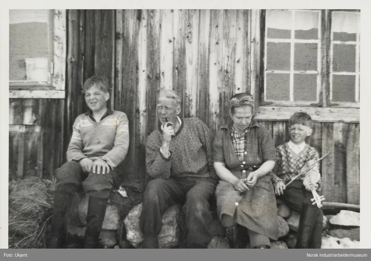 Familien Vågen sitter mot husveggen på gården Øst-Førnes på Møsstrond. Johan sitter ved siden av faren Olav som røyker en pipe. Olav jr. sitter ved siden av moren Sigrid med en fele