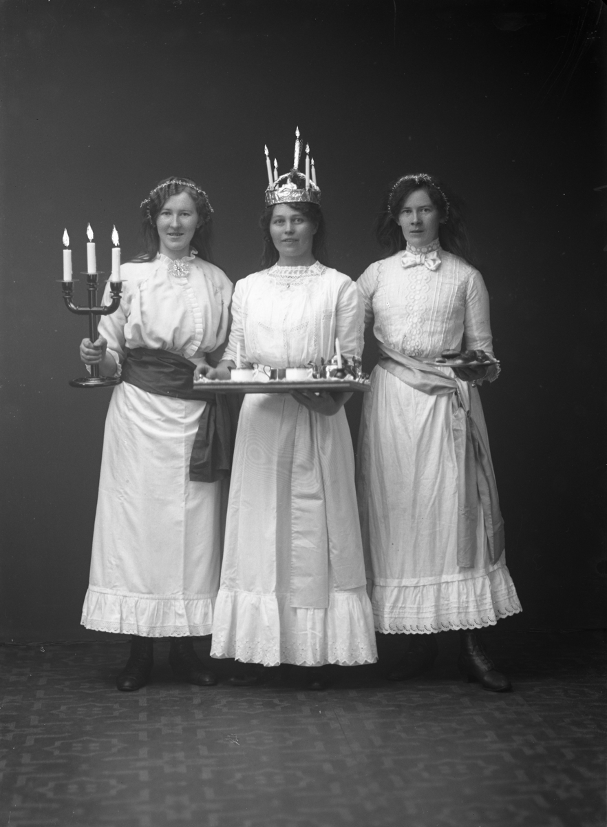Lucia med tärnor, ateljébild. Bilden föreställer Lucia med bricka samt två tärnor; en med ljusstake i höger hand och den andra med kakfat med lussebullar i höger hand.
Se även bild GB2_5558)