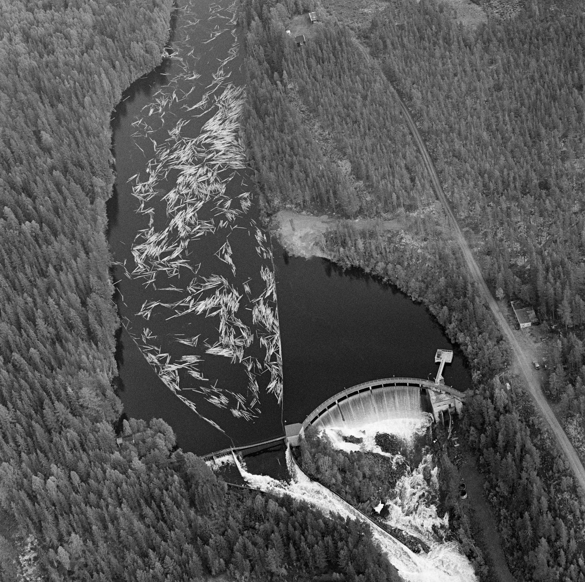 Flyfotografi tatt over kraftverksdammen i Osfallet i elva Søndre Osa i Åmot i Hedmark i mai 1984, den siste sesongen det foregikk tømmerfløting i denne delen av Glomma-vassdraget.  Bildet er tatt fra vest.  Fotografiet viser hvordan tømmerstokkene kom flytende med strømretningen over kraftverksdammen, der det var lagt ut lenser for å lede virket mot tømmerløpet som var plassert mot det nordre landet, hvor fallhøyden var mindre enn nedenfor den buete hoveddammen mot søndre landside.  Etter å ha passert dammen ble stokkene ført med strømmen i kvitskummende vann ned mot utløpet i elva Søndre Rena, cirka to kilometer lengre vest.  Damanlegget på fotografiet skal ha vært ferdigstilt i 1947.  Den ble noe ombygd i 1981.  Ettersom vassdragslovgivinga påla dem som drev tømmerfløting å fjerne installasjoner i vann ved opphør av virksomheten, ble tømmerløpet ved Osfallsdammen revet umiddelbart etter at fløtinga på dette fotografiet var avviklet.  Energiproduksjonen ved denne lokaliteten foregår i en kraftstasjonsbygning fra 1914, cirka en kilometer nedenfor den avbildete dammen, hvor det er installert to francisturbiner som kan yte 5.5 megawatt.  Den opprinnelige dammen ble ødelagt ved et dambrudd i 1916.  Det nåværende kraftverket utnytter et fall på 41 meter i elva Søndre Osa.  Vegen på sørsida av vassdraget fører ned til kraftstasjonen og derfra mot Fylkesveg 215 ved Oset.