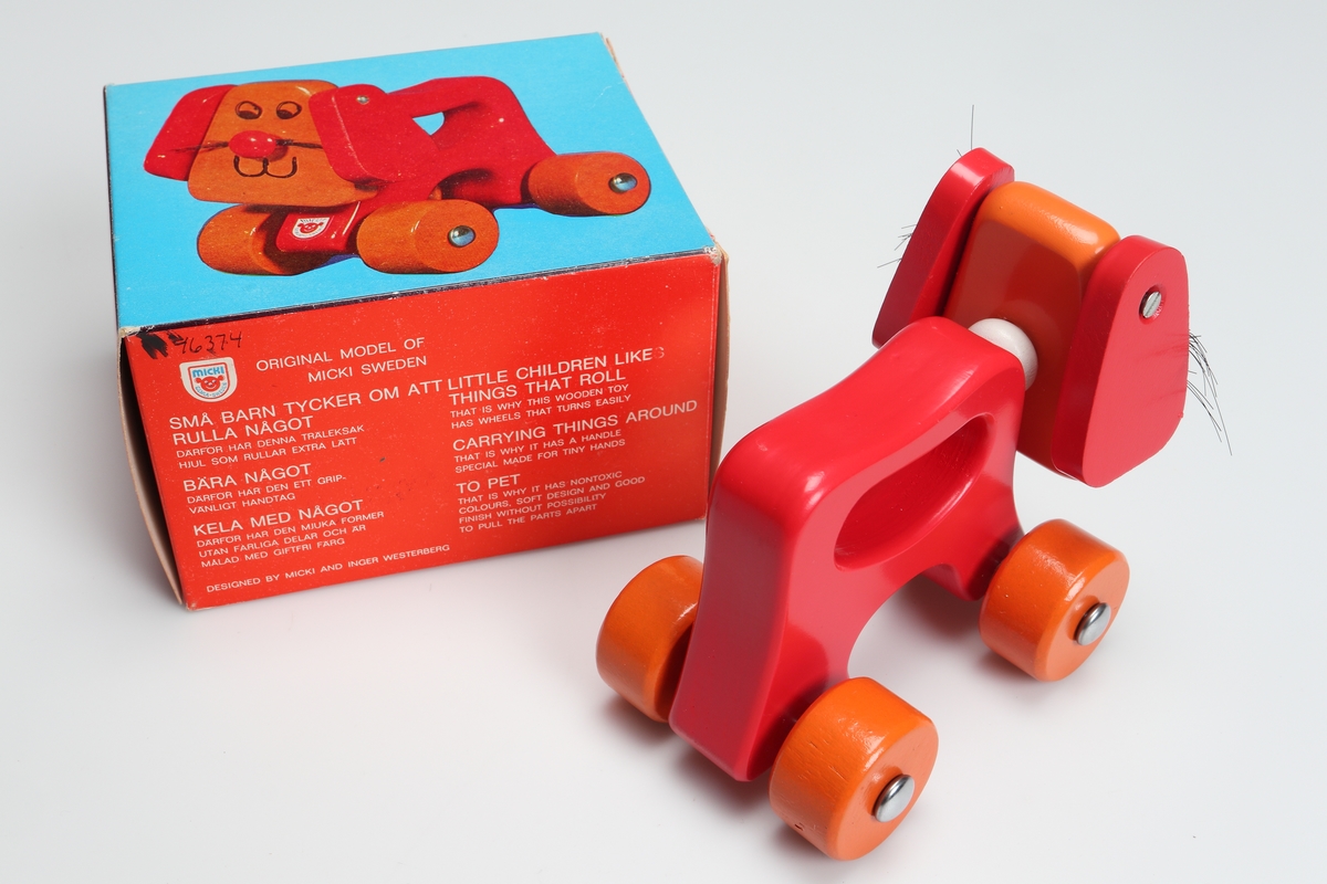 Rödlackerad leksakshund på fyra hjul. Ryggen är utformad som ett handtag. Hjulen är lackerade i orange färg. Morrhåren är gjorda av plast. Design: Inger Westerberg. Den är förpackad i en röd och blå pappkartong med bild och text på tyska, engelska och svenska.

Inskrivet i huvudkatalog 1982.