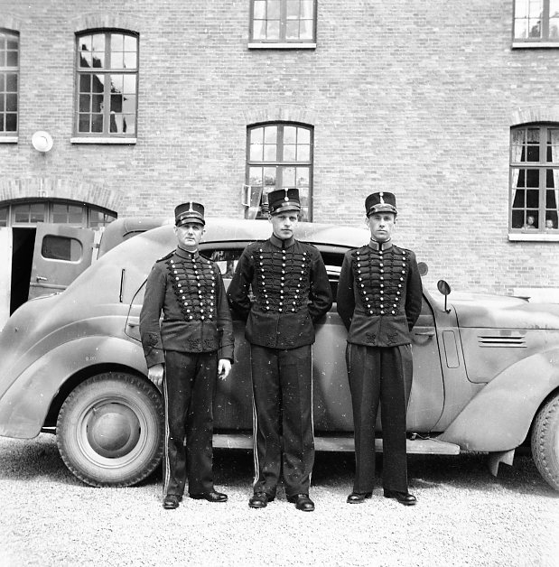 Underofficerare i uniform m/ä. Kaserngården A 6. Fordonet är en Volvo PV 53-56 och kamouflagemålningen säger att fotografiet är taget efter 1941.