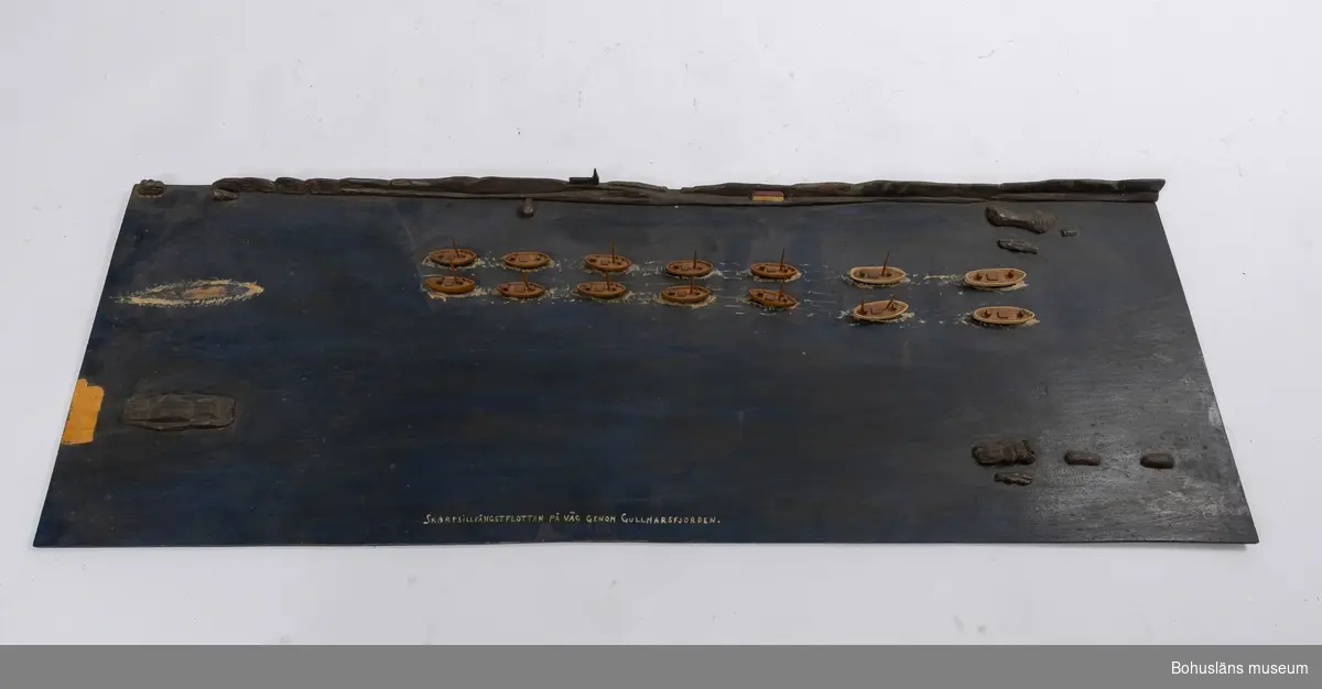 Rektangulär träplatta med fjorton båtar, två och två, i en rad.
I fonden är det en siluett av ett kustlandskap med berg, ett hus och en kyrka.
Text framtill på plattan: "SKARPSILLSFLOTTAN PÅ VÄG GENOM GULLMARSFJORDEN".
En större båt saknas. Master saknas på vissa av båtarna.

Ur handskrivna katalogen 1957-1958:
Skarpsillfångstflottan
Modeller på platta. Plattans mått: 116 x 42. Föremålen hela.
Från kapten Olssons saml., Fiskebäckskil.

För ytterligare information om förvärvet, se UM005087.