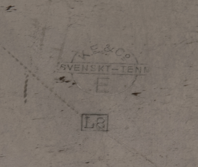 Minnestallrik av tenn med graverat motiv av Kiruna järnvägsstation i mitten. Motivet har en rektangulär ram. Tallriken har ett brett bräm indelat i flikar med en blomma i relief på varannan flik. Upphängningsögla fastlödd på baksidan.
