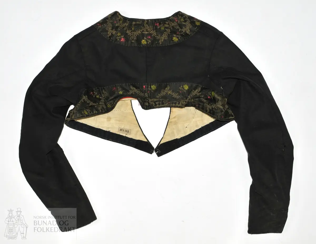 Kvinnetrøye i svart klede med fôr i ubleket bomull (toskaft). Trøya er kanta med et 7 cm bredt svart silkeband med mønster i rødt, grønt og brunt. Nederst på ermene 7 cm splitt. Sjå skisse.