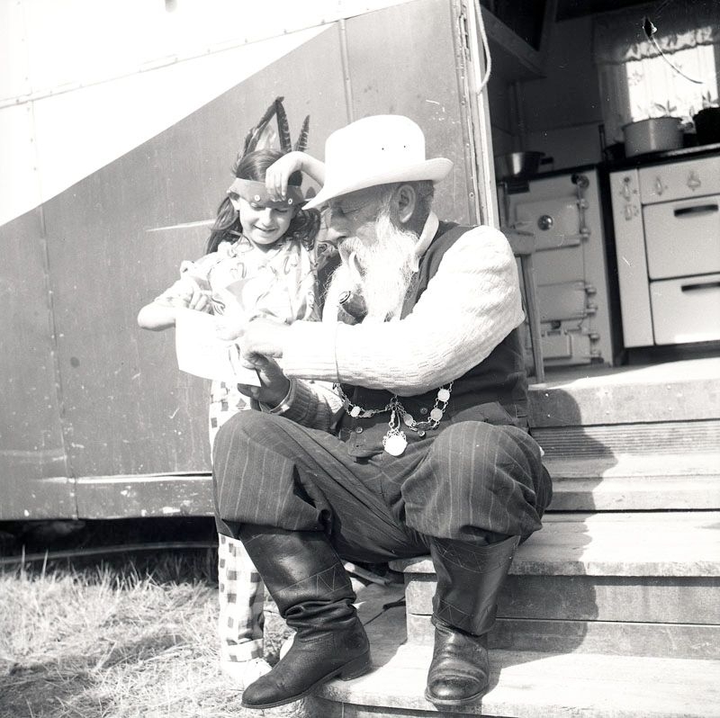 Fotografiet är taget i juli 1952 i samband med skolundervisning i det romska lägret i Ekeby, ca 4 km väster om Eskilstuna. Ett barn med fjädrar i håret tittar på ett papper tillsammans med en äldre man.