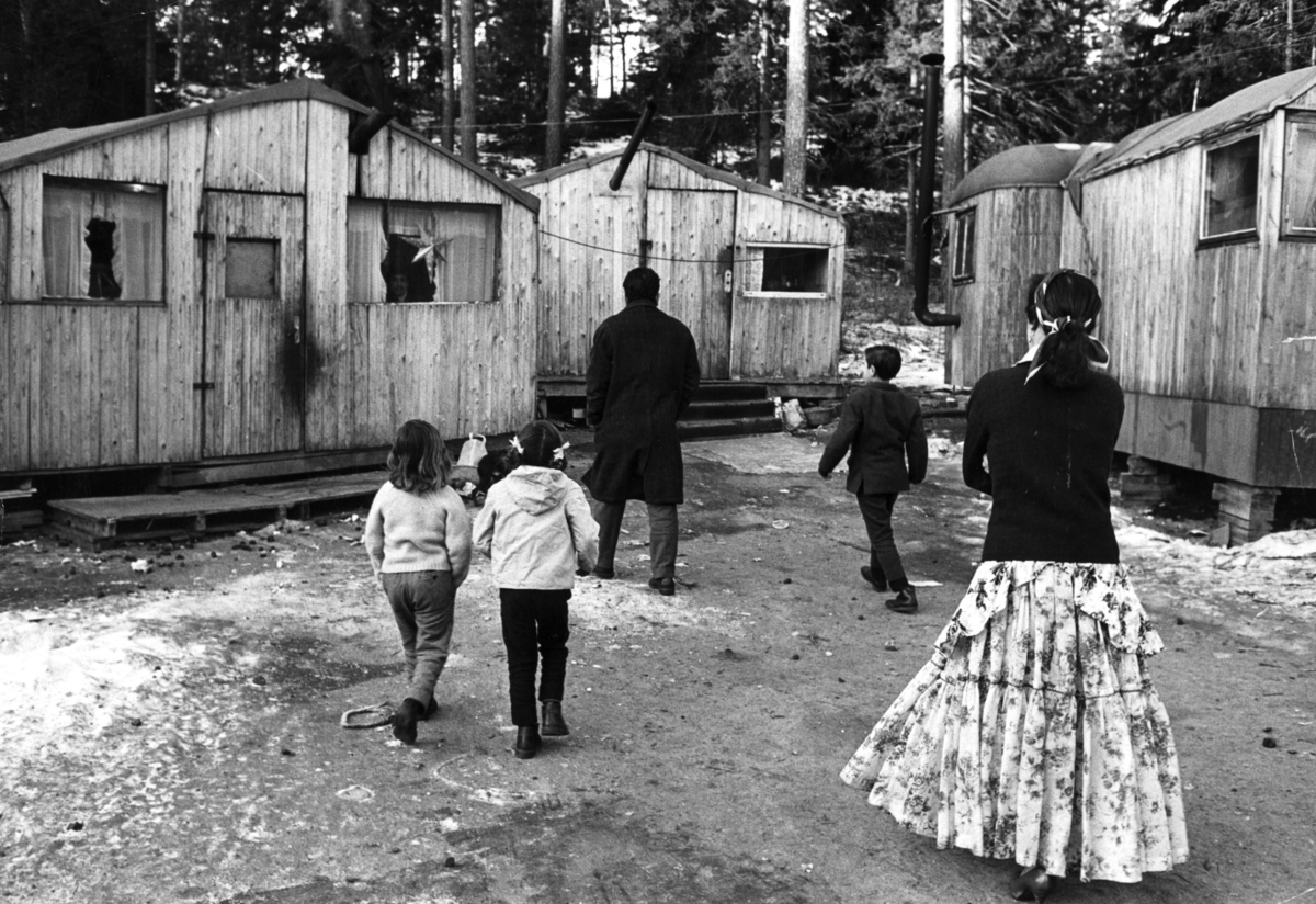 En tillfällig lägerplats för svenska romer med baracker och bostadsvagnar utanför Stockholm. Över "gården" mellan vagnar och stugor går tre barn samt en man och en kvinna med ryggarna vända mot kameran.