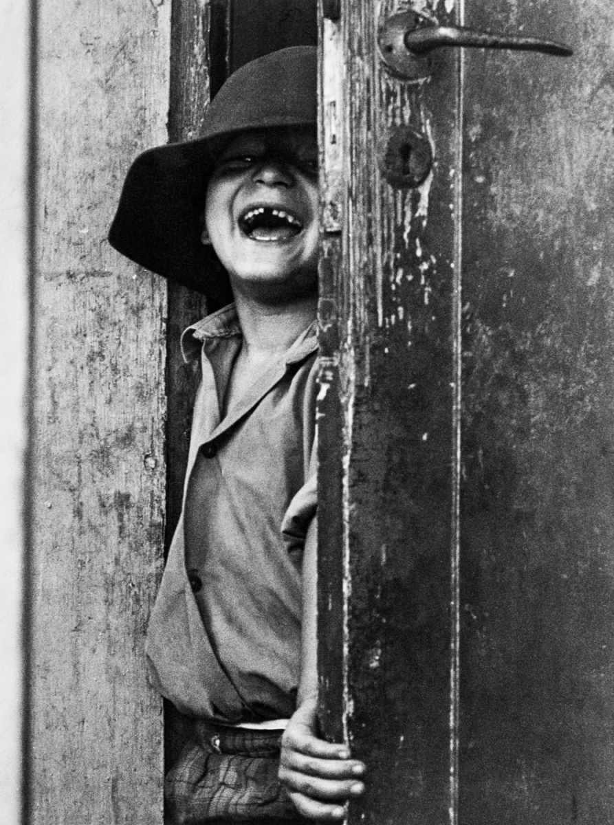 En pojke står i en dörröppning och ler mot kameran. Han bär en hatt.