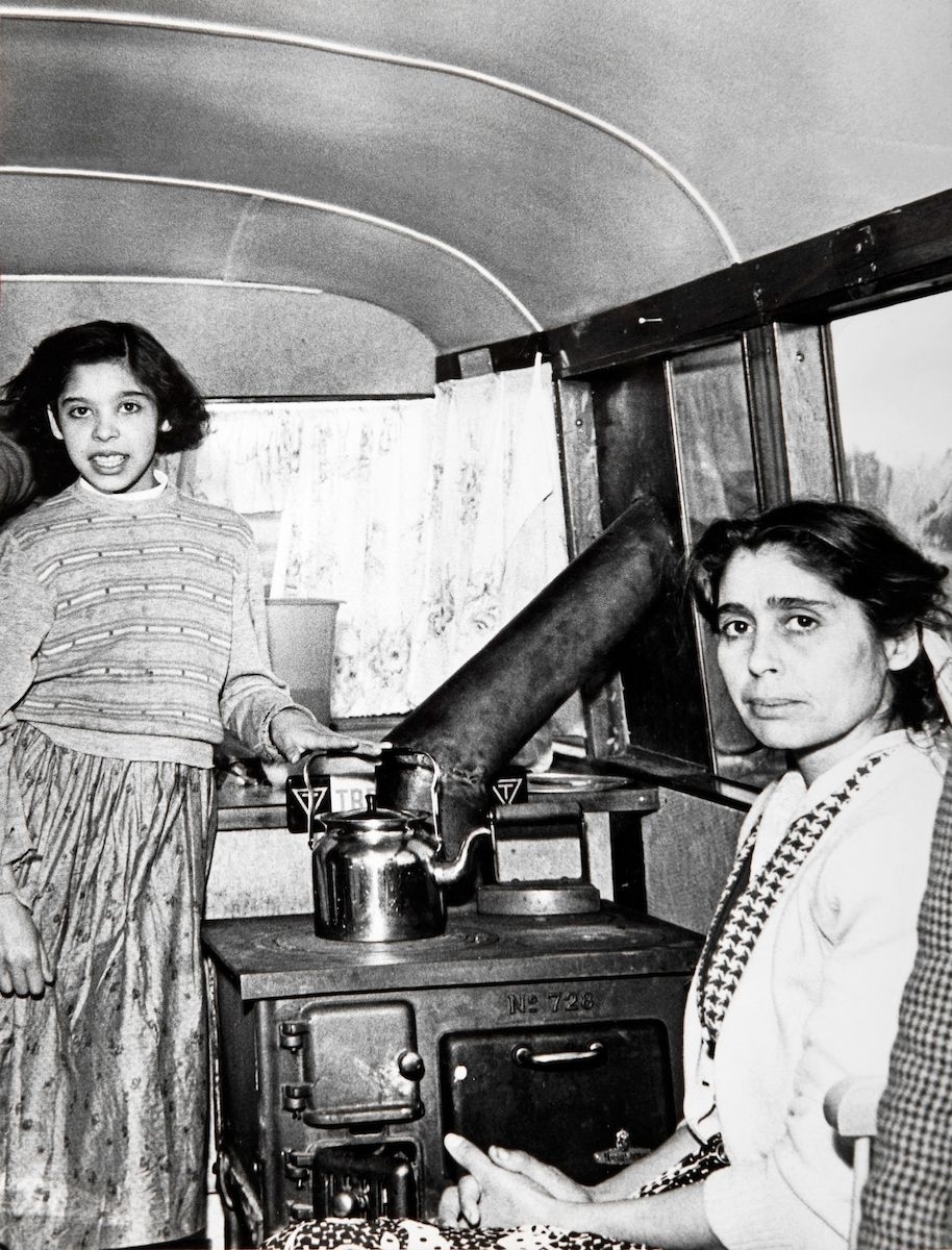 Bilden föreställer mor och dotter i sitt kök. Köket finns i en buss, som invånare i Önum och Vara tillsammans ordnat fram som bostadsvagn för den romska familjen, som tidigare bodde i tält. Flickan kokar något i en kanna på en spis.