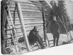 En gruppe menn står og sitter utenfor en tømmerbygning. Ski 