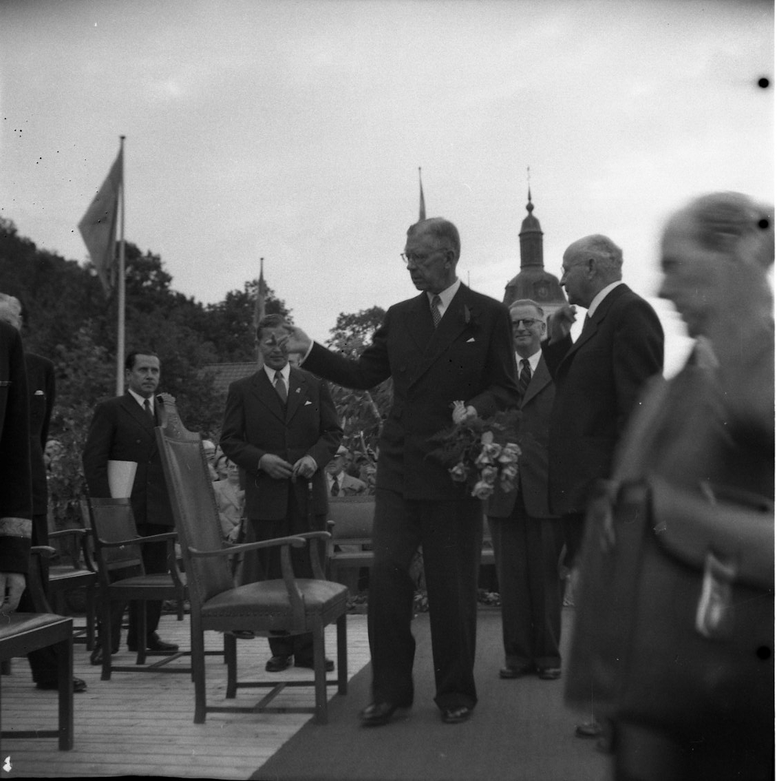 Eriksgata 1954. Kung Gustaf VI Adolf står på en särskilt uppbyggd estrad i Braheparken med en rosbukett i handen, han gestikulerar. Omgiven av män, bl a stadsfullmäktige vice ordf Ture Ekvall och ordf Ivar Ekvall som står till höger.