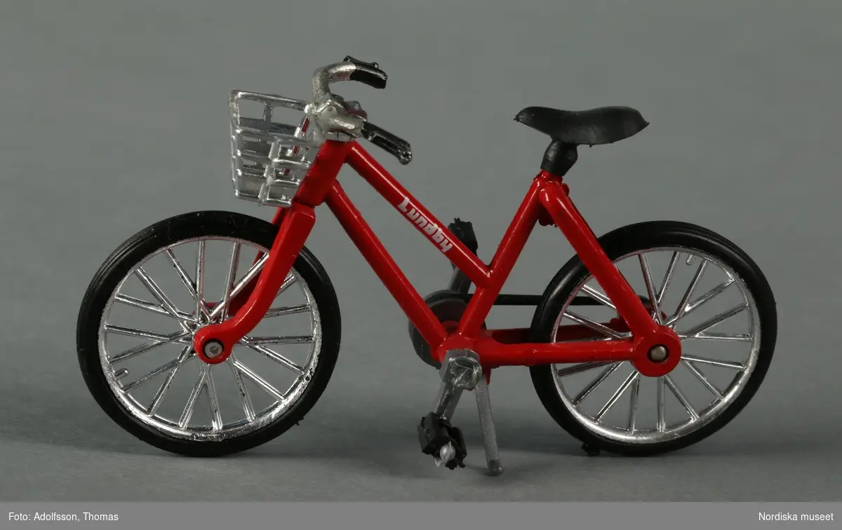 En röd flickcykel märkt "Lundby". Den rödmålade stommen är av metall, medan sadel, hjul, cykelkorg, pedaler, kedja, handtag mm är av plast. Pedalerna, hjulen och stödet är rörliga. Cykeln hör till dockskåpsinredningen i garaget i källarvåningen i dockskåp NM.0331721+.