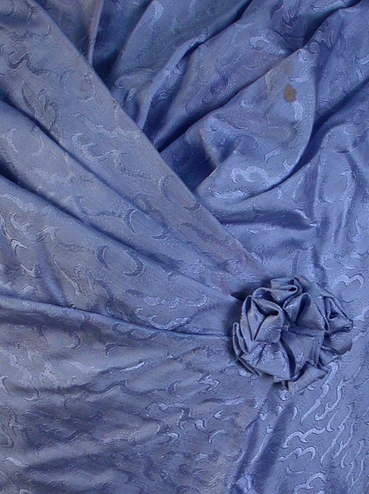 Mönstrad ljusblå sidenklänning, tvådelad. 
1) Liv: 2 våder fram, 1 bak, med kil i var sida. 4 st lagda veck från ärmhål, draperade mot brösthöjd, avslutad med sydd tygros som fästes med hyska och hake. Våderna sydda på vitt maskinsytt bomullsfoder med 9 st insydda sprialfjädrar. Fodret knäppes med 13 st hyskor och hakar. 3 st hakar i ryggen för fastsättning av kjol. Rund bandkantad hals med lös påsydd del som bildar ståkragen, fäst med 3 st hyskor och hakar. Smal isydd och avsmalnande tvåsömsärm, lite rynkad vid armbåge, svängd nederkant. Kort puffärm med mycket rynk i ärmhål och nederkant ovanpå den avsmalnande ärmen. Puffärmen kantad med 2,5 cm styv linning av samma tyg. Isatt tylldel i båda puffärmar för mer volym. Ärmen helfodrad. Ryggvåden slät och något snedsydd. 
Mått: längd 43 cm
2) Kjol: 5 st utställda våder. Helfodrad med vott bomullstyg. Litet släp. 2 st nedsydda veck framtill. Bandkantad med samma tyg i linningen. Knäppes med 2 st hyskor och hakar mitt bak, isydd ficka i sprundet mitt bak. Vid sprundets början 10 cm rynk. 11,5 cm bred tyllbård med spets i nederkant, fastsydd i fodret i kjolens nederkant. 2 st hällor fastsydda innanför linningen för upphängning.
Mått: längd 104-120cm.
3) Lös ståkrage: av samma tyg som klänningen. Spetsformad i ena kanten, rak nederkant och vågformad överkant. Fodrad med vitt silkestyg, insydda förstärkningar i fodret. I överkant veckad tyllspets. Påsydd blommönstrad spetsbård i överkant. Knäppes med hake och sydd hyska.
Mått: längd 41cm, bredd 5-7,5 cm.
4) Midjeband: Vågformat. I samma tyg som klänningen. 4 st spiraler insydda i fodret. Rynkat avslut i ena kanten och volangrynk i andra kanten. Knäppes med 3 st hakar och sydda hällor. 
Mått: längd 73 cm, bredd 9-11 cm.
