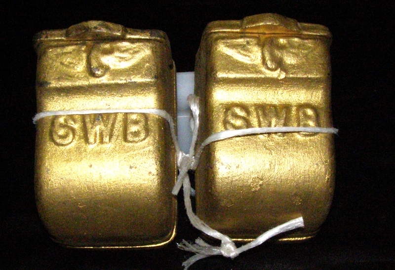 Två askkoppar i gjutgods med guldfärgad lackering. Med lock, och avsedd för upphängning på vägg. Dekorerad med okrönt vinghjul samt initialerna SWB som står för Stockholm-Westerås-Bergslagernas Järnväg.