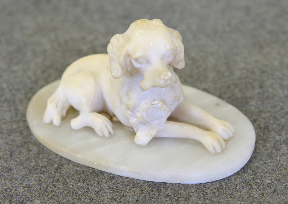 
Prydnadsfigur i form av en liggande hund. Tillverkad av vitt, oglaserat gods monterad på en oval platta av marmor.