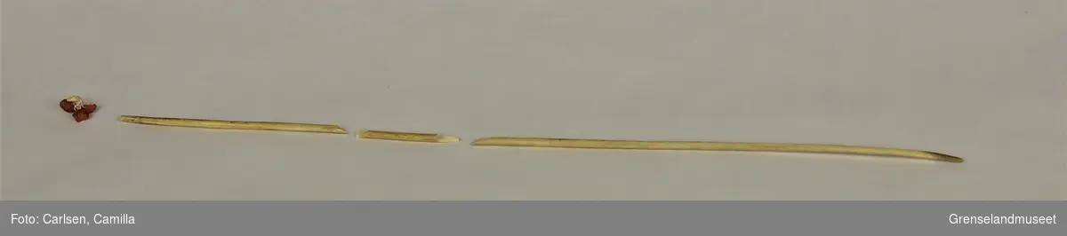 Et par lange strikkepinner av bein, med røde endeknopper. Den ene pinnen har brukket i tre deler. Endeknoppen på denne har løsnet og er i tre deler. 