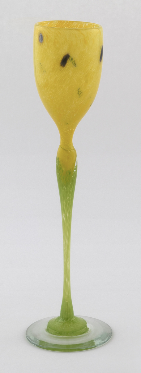 Høyt drikkeglass med tulipanlignede utforming. Eggeformet kupa i gult halvgjennomskinnelig glass med sorte prikker. Stetten er utført i lysegrønt opakt glass, som hviler på en transparent fot.