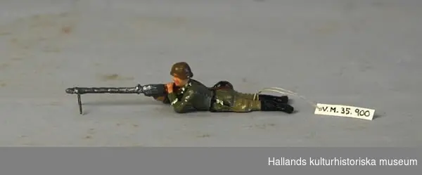 Leksakssoldat av trämassa på ståltrådsskelett i tysk uniform. Soldaten ligger raklång och skjuter med en kulspruta. Målad i grönt, brunt, svart, grått och skärt.