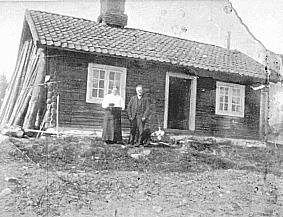 Bildet viser to mennesker som står foran et lite hus. Bildet er i dårlig kvalitet og svart/hvitt.