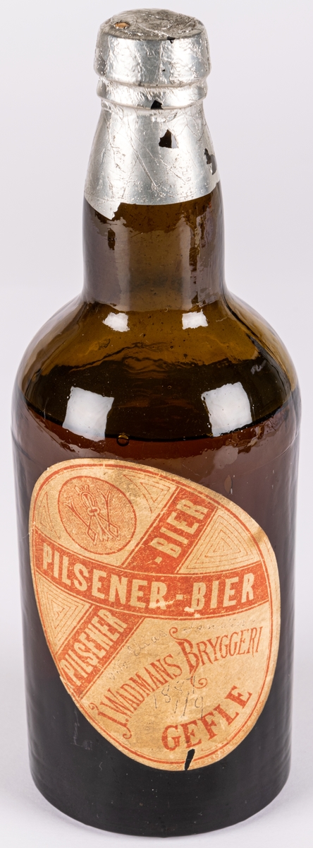 Pilsnerflaska med innehåll, enligt etiketter: Pilsener-Bier. Från J. Wadmans bryggeri, Gefle.
Brunt glas, folie över kork. Etikett av papper.