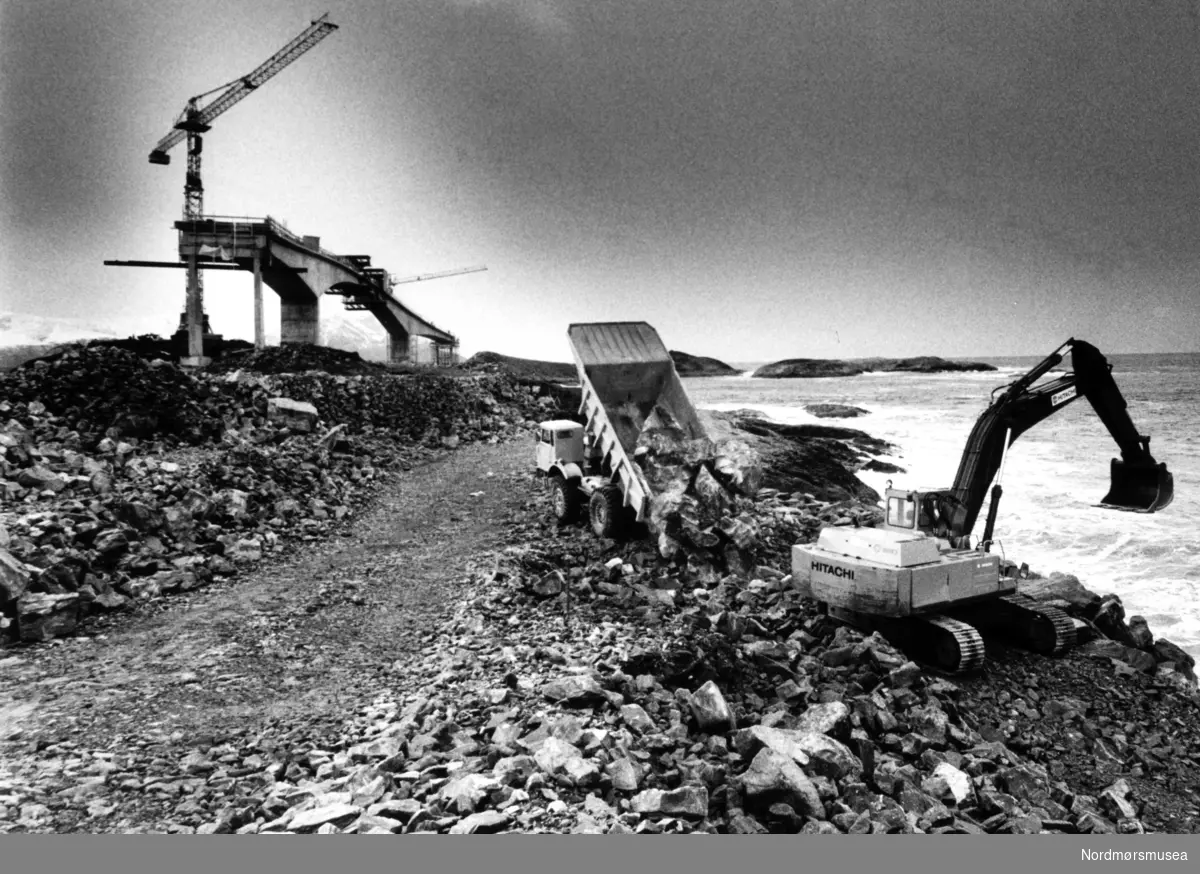 Fra byggingen av Atlanterhavsveien mellom Averøy og Eide i Møre og Romsdal. En veistrekning på 8.274 meter. Anlegget består av åtte broer på til sammen 891 meter og går over holmer og skjær. Veien kostet 122 millioner i 1989 og var delvis bompengefinansiert. Anlegget ble åpnet 7. juli 1989 og bommen ble fjernet i juni 1999. Atlanterhavsveien er landets åttende best besøkte naturbaserte turistattraksjon (2004) med 283.500 besøkende. Veistrekningen er foreslått vernet i "Nasjonal verneplan for veger, bruer og vegrelaterte kulturminner." (Kilde: Wikipedia Norge). Bildet er fra avisa Tidens Krav sitt arkiv i tidsrommet 1970-1994. Bildet er fra avisa Tidens Krav sitt arkiv i tidsrommet 1970-1994. Nå i Nordmøre museums fotosamling.