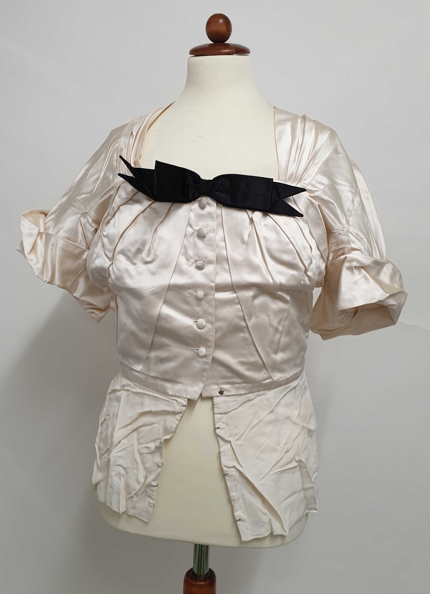 Eggeskall-farget bluse av silke, med svart sløyfe i fronten, breder ermer med oppbrett, seks knapper i front. Nedenfor blusen er det sydd fast et slags underskjørt i tynt stoff, med henper.