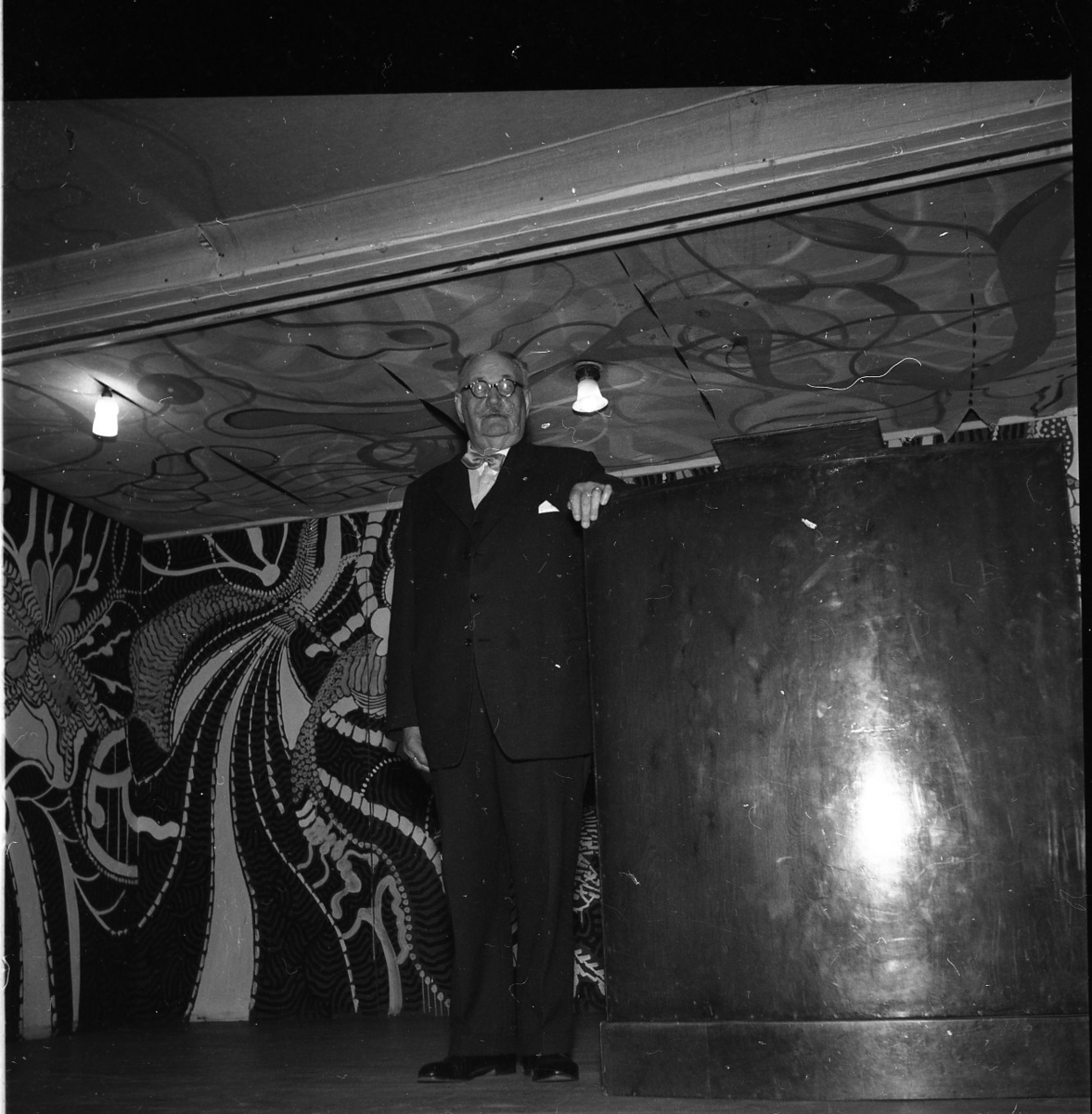 En äldre man i kostym står vid en talarstol på en scen med 60-tals dekor. Han håller sannolikt ett tal.