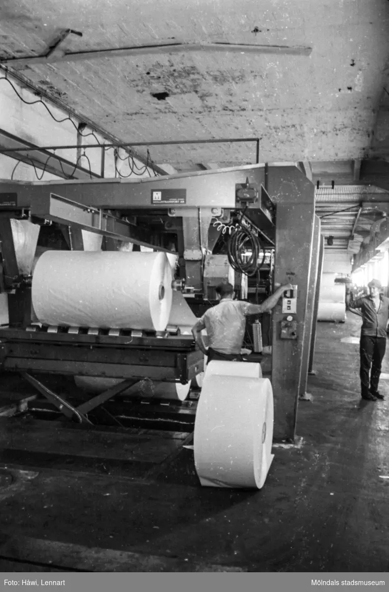 Män i arbete vid maskin. Antagligen Voldemar Mei med ryggen vänd mot kameran. Rullar läggs i klippen och klippta ark kommer ut på andra sidan av maskinen. Klipp 5 (fabrikat Masson/Mason & Scott), byggnad 10. Pappersbruket Papyrus i Mölndal, hösten 1970.