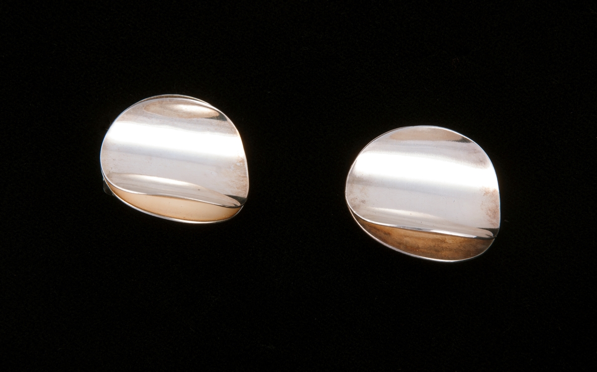 Består av to like store sirkulære sølvringer som er satt sammen langs en midtakse. På hver siden av aksen er ringene bøyd oppover i ulik grad, symmetrisk på hver side.