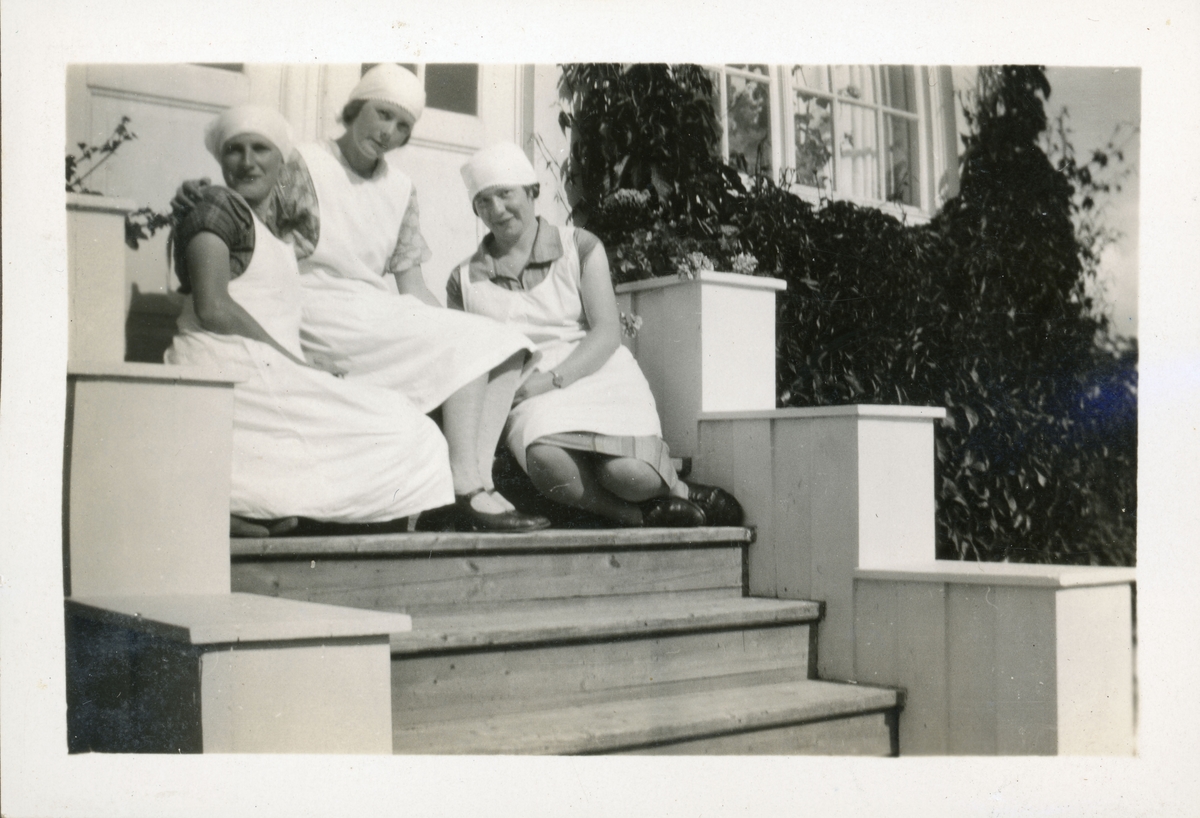 Elever fra husmorskolen på Skansgården, Kongsvinger, 1928. Tre unge kvinner med forklær og tørklær sittende i steintrapp foran huset.