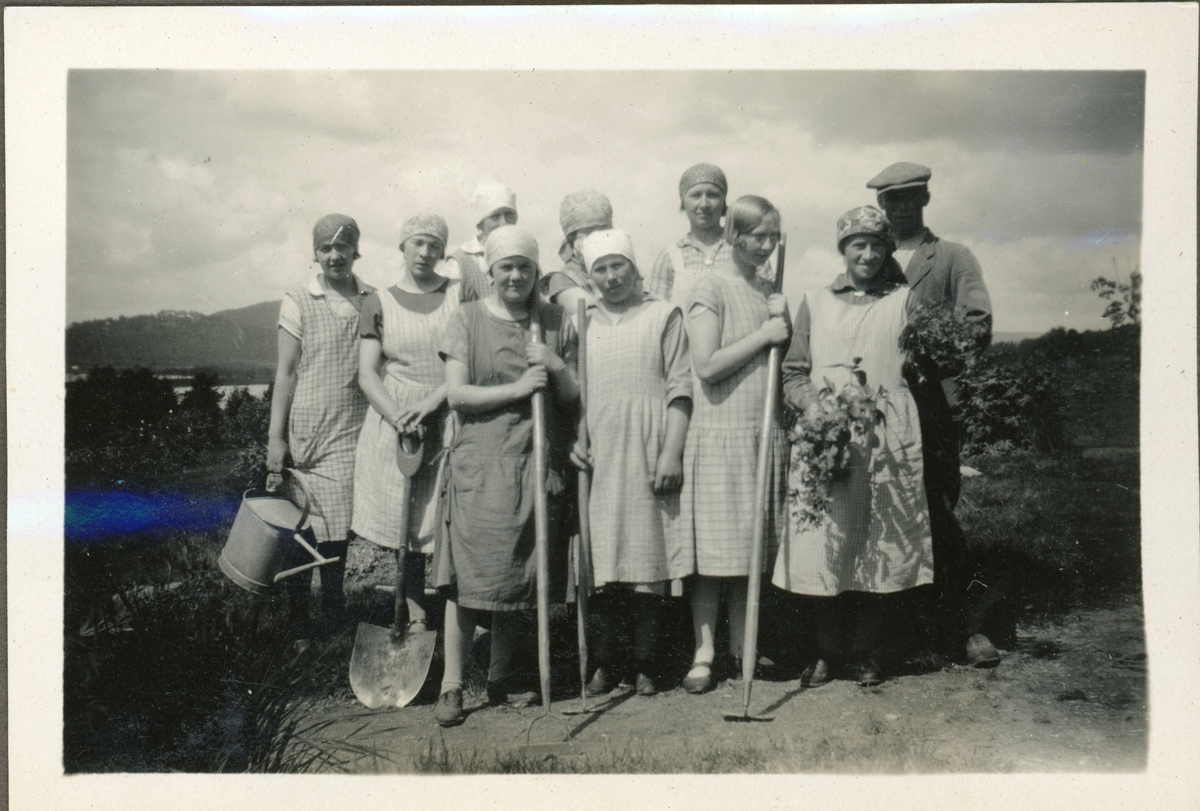 Elever fra husmorskolen på Skansgården, Kongsvinger, 1928. Ni unge kvinner og en mann i arbeidstøy med hageutstyr. Kvinnene er ikledd Skansgårdens vaskehabitt.