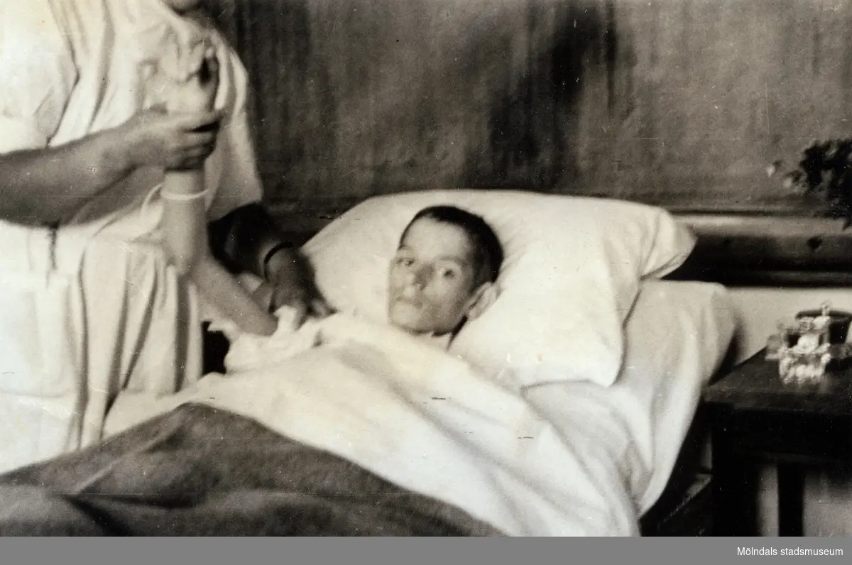 "Karantänsförläggning" på Kvarnbyskolan för överlevande från koncentrationsläger i Tyskland och Polen 1945.  
Skolan fungerade under denna sommar som beredsskapssjukhus.

En sköterska håller upp armen på en ung man som ligger i en sjuksäng.