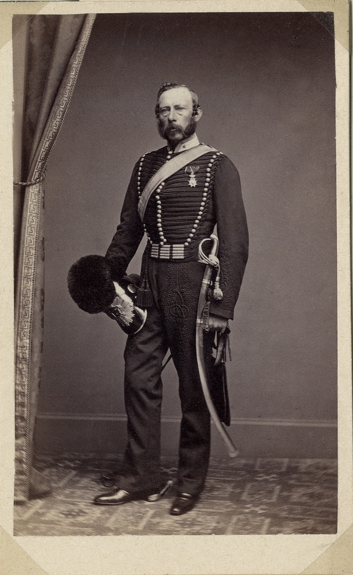 Porträttfoto av en medelålders man i militär paraduniform med kask och sabel m.m.. Till vänster syns ett draperi. 
Helfigur. Ateljéfoto.