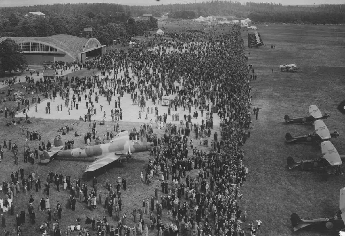 Flygbild av flygfältet på F 3 Malmen vid flygdagen Flygets dag, sommaren 1938. Publik och flygplan. Flygplan B 3 Junkers Ju 86K till vänster, S 6 Fokker C.V. till höger, både i förgrund och längre upp. I mitten till höger P 1 Sparmann S 1-A och Sk 11 Tiger Moth.