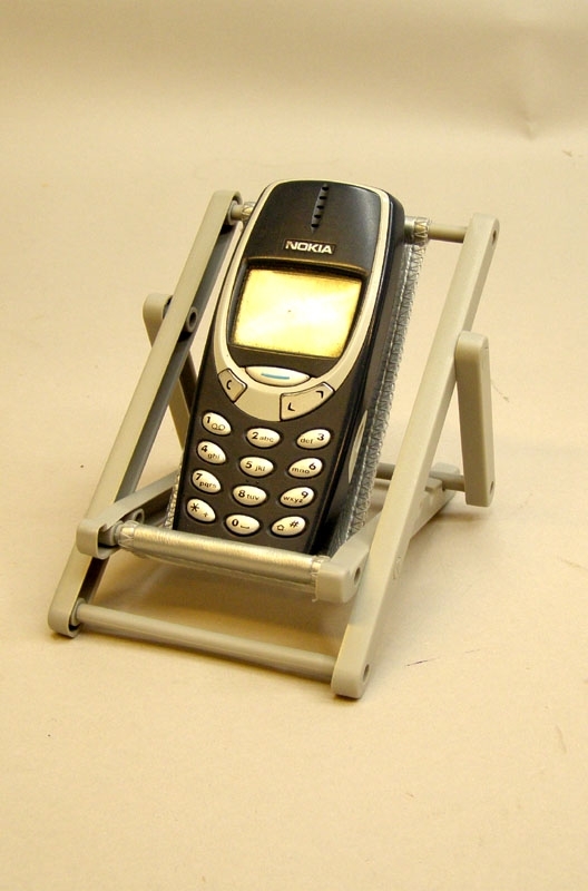 Bordsställ för mobiltelefon, i form av en solstol i miniatyr, grå ställning med ställbart läge.
Dynan är silverfärgad med röd logga för Tåg i Bergslagen.