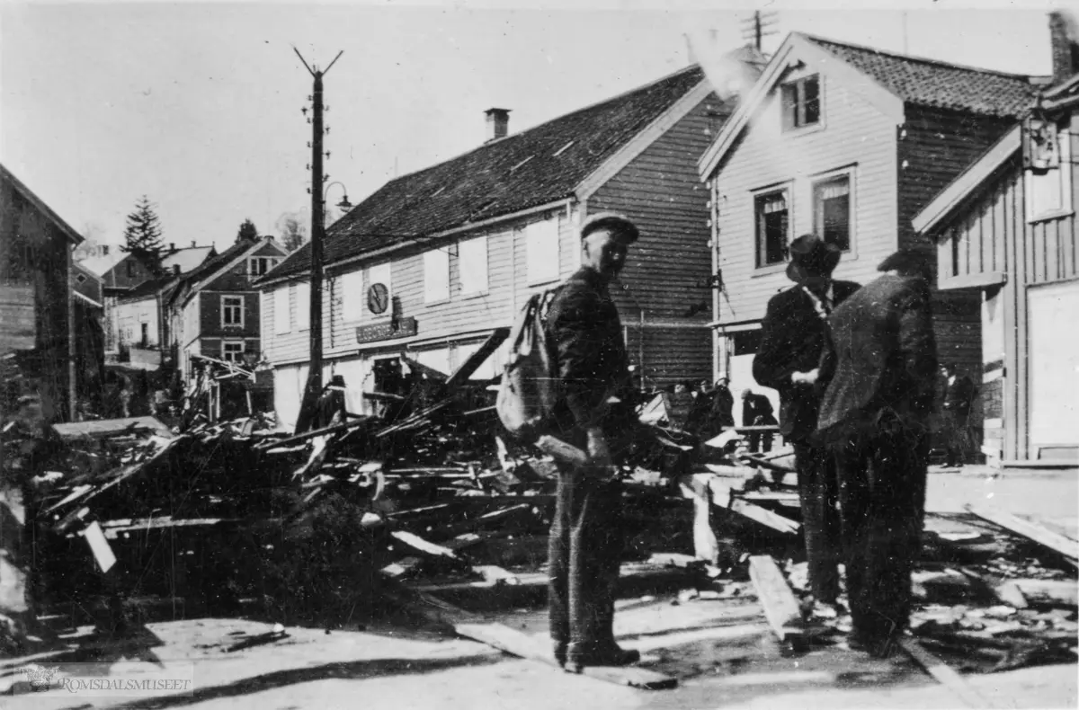 Dette bildet er tatt etter bombingen av den østlige del av S.Petterssons bygg ved Torget torsdag 25.04.1940. .Fra høyre ser en et hjørne av "Høstmarkgården", O.Løckens foretningsgård, Dahls bokhandel og gårdene oppover Myrabakken med Thommassens gård i forgrunnen.