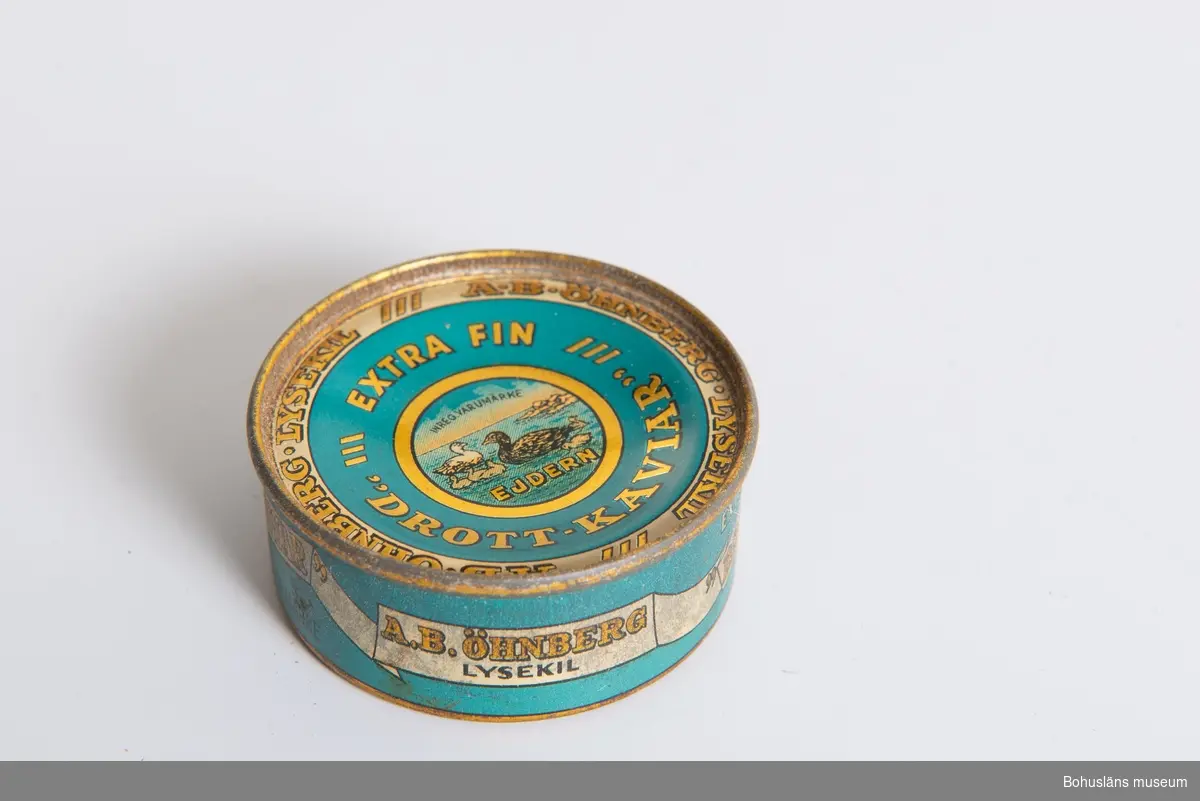 Konservburk för: "Extra fin drott-kaviar". Burk i turkos, guld och beige färg. 
Motiv med ejdrar (av varumärket ejdern).