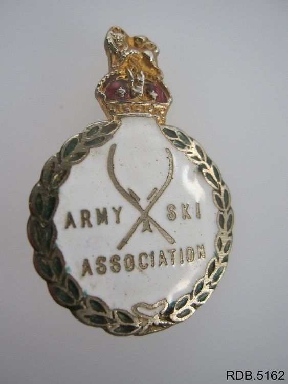 Rund pins med ei kongekrone på toppen. Ei nål på baksiden. Army Ski Associations logo på forsiden.