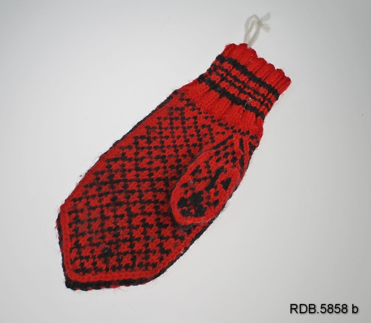 En håndstrikket barnevott i ull. Rød bunn med svart mønster: 2 1/2 åttebladrose på håndbaken og rutemønster i handflata. Vrangbord  2 r og 2 vr. med 4 striper.