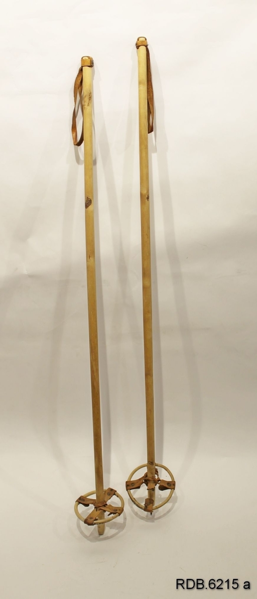 Et par skistaver av tre med brune, fastklinkede lærhemper og bambustrinse med brunt lærkryss. Jernpiggen på stav A er borte.