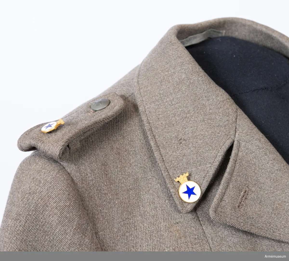 Jacka sydd i grå yllediagonal med märken och knappar för Svenska Blå Stjärnan. Till jackan hör ett skärp i samma tyg.