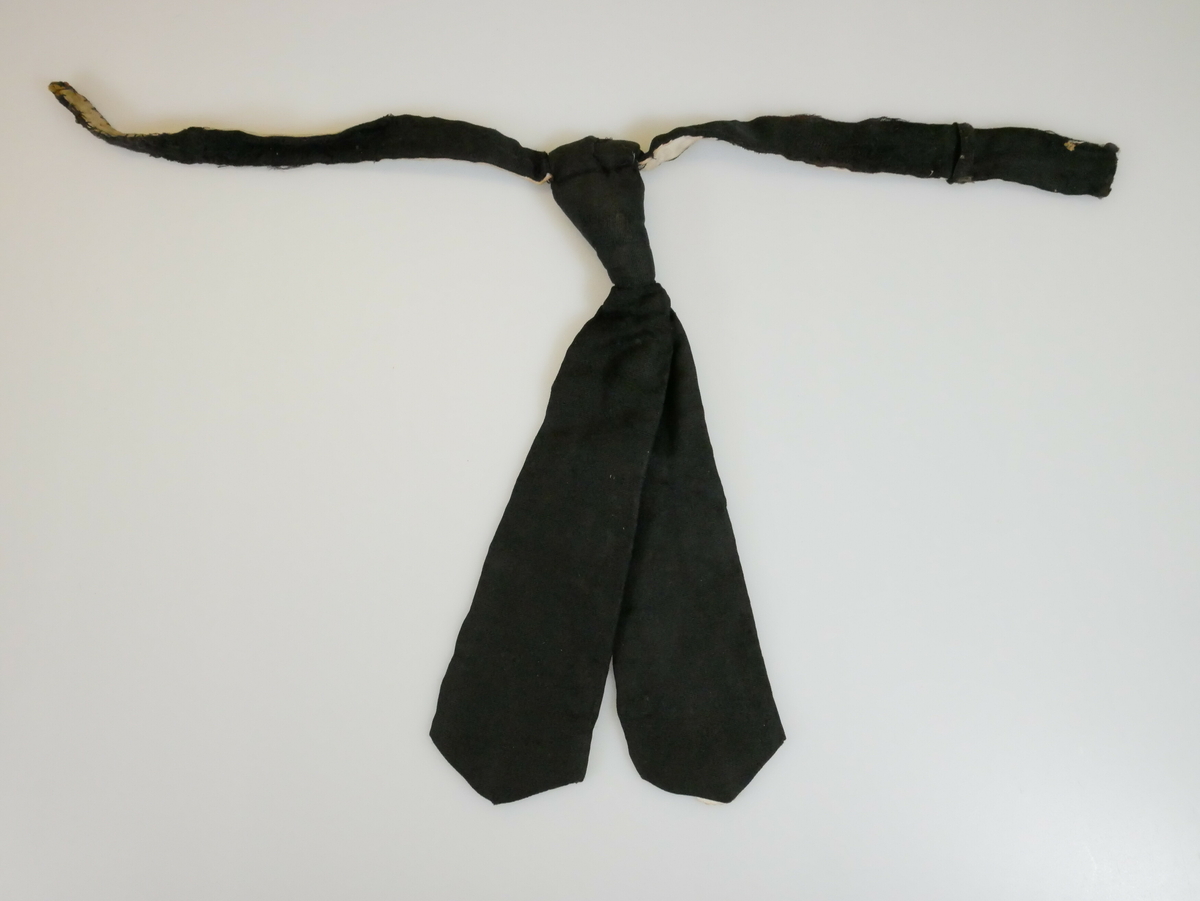 Kort svart slips. Slipsknyten er sydd sammen. De to slipslengdene er like lange og spisse nederst. De er sydd sammen på midten med en søm. På baksiden er det et hvitt for. En liten rund hvit papirlapp er sydd på bak nederst på den ene slipslengden. 